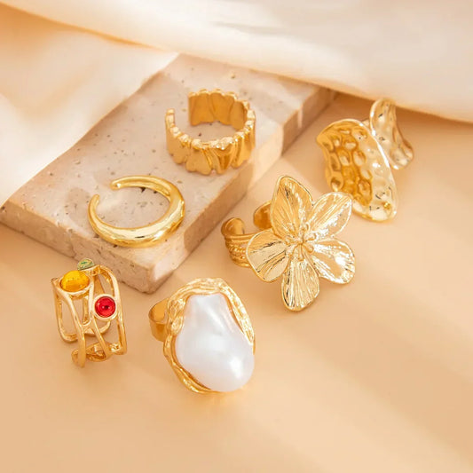 Assorted gold statement rings with floral, pearl, geometric, and textured designs on beige background