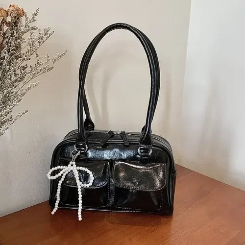 Shiny black handbag with dual handles, front pockets, and pearl bow charm on wooden table